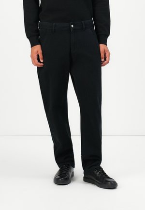 Mann iført svarte slim-fit bukser, svarte snørebåndssko og en svart langermet topp mot en hvit bakgrunn.