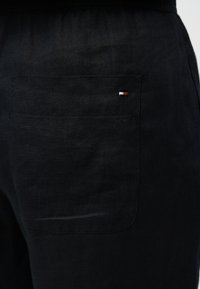 Κουστούμι από μαύρο ύφασμα σε κοντινή λήψη που δείχνει μια πίσω τσέπη με ένα μικρό ορθογώνιο λογότυπο σε κόκκινο, λευκό και μπλε κοντά στην πάνω δεξιά γωνία.