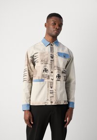 Helle beige Jacke mit Denim-Kragen, gemischten Mustern, zwei großen Vordertaschen und knopflochverschluss. Text- und grafische Details sichtbar.