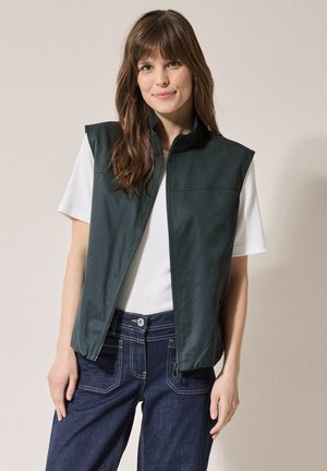 Mörkgrön ärmlös väst med dragkedja och bröstficka, bärs över en vit skjorta, matchad med mörka jeans. Slät textur.