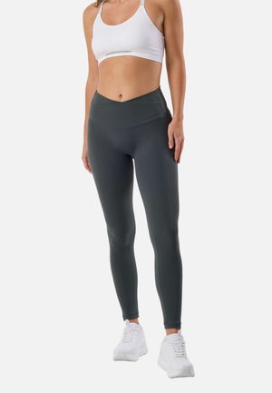 Kobieta ubrana w biały sportowy stanik, ciemnoszare legginsy z wysokim stanem oraz białe buty sportowe, stojąca na jednolitym tle.