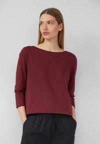 Burgunderfarbenes Langarmoberteil mit weitem Ausschnitt und strukturierter Stoffqualität, kombiniert mit schwarzen Stretchhosen mit elastischem Bund. Lässiges Outfit-Design.