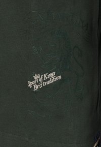 Tummanvihreä puuvillainen T-paita, jossa on kirjailtu leijonakuosi ja valkoinen teksti "Sport of Kings Brit tradition." Hienovarainen tekstuuri.