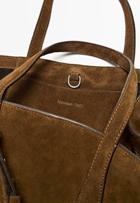 Intérieur d'un sac fourre-tout en daim marron avec des accessoires en métal, un logo embossé et des coutures robustes. Les poignées s'étendent du haut pour un transport facile.