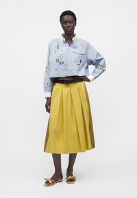 Mujer con una camisa azul clara corta con botones y parches de colores, falda midi amarilla plisada, cinturón marrón y sandalias marrones con nudos.