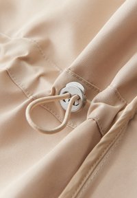 Tessuto beige con una texture, caratterizzato da un occhiello in metallo e un anello in corda beige. Il design include dettagli arricciati lungo il bordo.