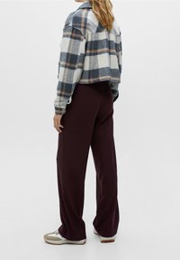 Femme debout, vue de dos, portant une veste à carreaux courte, un pantalon large violet foncé et des baskets blanches, sur un fond uni.