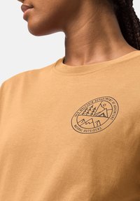 Beżowy bawełniany t-shirt z okrągłym dekoltem, na którym znajduje się czarna okrągła grafika przedstawiająca góry i drzewa, opisana jako "Departament Przygody".