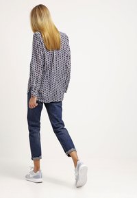 En blond person går bortåt iförd en mönstrad långärmad skjorta, uppkavlade mörka jeans och vita sportskor.