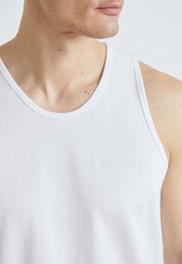 Balts kokvilnas topiņš bez piedurknēm, ar plašu dekoltē un diskrētu izspiestu logotipu "KARL LAGERFELD" uz krūškurvja. Gluda tekstūra.
