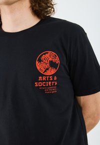 Black cotton t-shirt featuring a red graphic design with a wave and text reading "ARTS & SOCIETY" with a small tagline underneath.