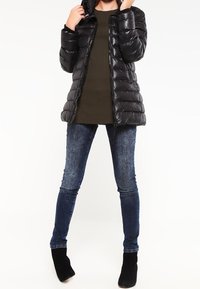 Chaqueta acolchada negra con capucha, con un acabado brillante, cierre de cremallera y diseño acolchado. Combinada con jeans de mezclilla azul oscuro y botines negros.