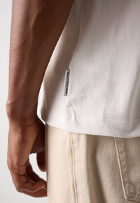 White cotton t-shirt with a side seam label, paired with light beige trousers featuring diagonal ribbed texture and visible stitching.