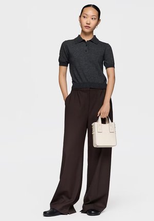 Mujer con camiseta polo de manga corta gris oscuro y pantalones marrones de pierna ancha, sosteniendo un pequeño bolso blanco con ambas manos.