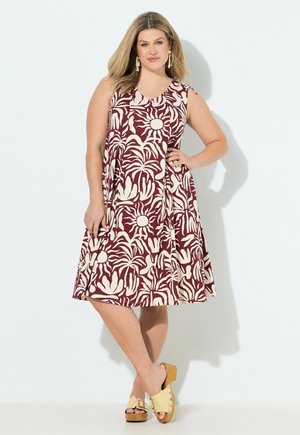 Kvinde i ærmeløs burgunderrød kjole med beige abstrakt blomsterprint, stående med hånd på hoften, iført beige platform sandal med åben tå.