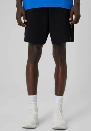 Mężczyzna w czarnych szortach, białych skarpetkach i białych butach sportowych, stojący na jednolitym jasnym tle.