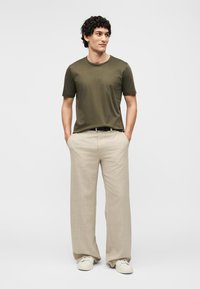 Άνδρας με κατσαρά μαλλιά που φοράει μια ελιάς πράσινο μπλουζάκι, μπεζ παντελόνια με φαρδιά πόδια και λευκά αθλητικά παπούτσια, που στέκεται με τα χέρια στις τσέπες σε απλό φόντο.