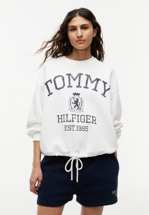 Vrouw met lang haar die een wit Tommy Hilfiger sweatshirt en zwarte shorts met trekkoord draagt, staand tegen een effen achtergrond.