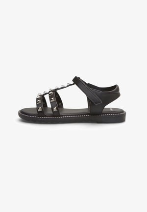 Schwarze flache Sandale mit drei genieteten Riemen über dem Fuß und verstellbarem Knöchelriemen auf weißem Hintergrund.