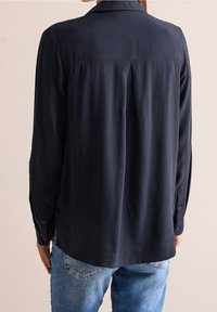 Chemise à manches longues bleu marine avec une surface texturée, des poignets à boutons et un pli dans le dos. Portée avec un jean en denim bleu clair.