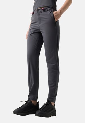 Person wearing dark gray slim-fit pants with zippered side pockets and black athletic shoes, standing with one hand in pocket.