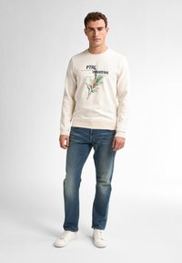 Crèmekleurig sweatshirt met een grafische afbeelding en de tekst "PTRL Industries," gecombineerd met blauwe jeans en witte sneakers. Simpele, casual outfit.