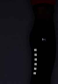 Černé sportovní legíny mají reflexní prvky po stranách, které zvýrazňují jejich elegantní design a přiléhavý střih. Logo Under Armour je viditelné.