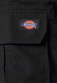 Shorts en tissu noir avec une grande poche avant. Un écusson en logo « Dickies » multicolore est montré de manière proéminente sur la poche.