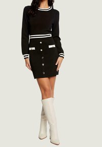 Pull noir en maille avec des rayures blanches au niveau du col et des poignets, associé à une jupe noire ornée de boutons dorés et d'accents de poches blancs. Bottes blanches montant jusqu'aux genoux.