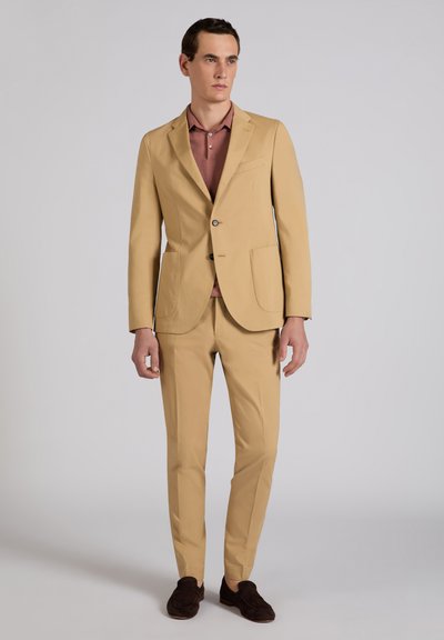 Homme portant un costume beige avec une chemise marron et des mocassins marron foncé, debout devant un fond gris uni.