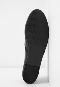 Semelle d'une chaussure noire avec un motif de grip texturé et la taille 37 embossée près de la voûte plantaire, positionnée debout contre des murs blancs en angle.
