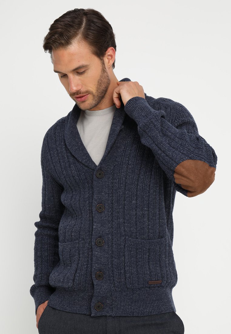 Navyblå stickad cardigan med ribbad struktur, knäppning, två fickor och bruna armbågsfläckar. Har en sjalkrage-design.