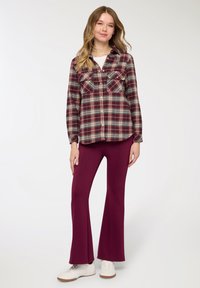 Camicia di flanella a quadri in bordeaux, beige e verde con doppie tasche sul petto; abbinata a pantaloni svasati bordeaux e sneakers bianche.
