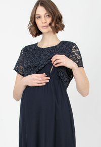 Marineblaues Umstandskleid mit einem Spitzenoberteil mit floralem Muster und kurzen Ärmeln. Es enthält eine diskrete Öffnung für den Stillzugang.