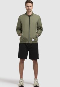 Olivgrüne Bomberjacke mit Reißverschluss, gerippten Bündchen und Saum. Verfügt über Seitentaschen und ein Logolabel am Saum. Getragen über schwarzen Shorts.
