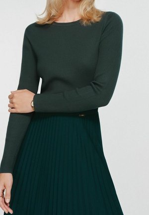 Femme portant un haut à manches longues côtelé vert foncé et une jupe plissée assortie, avec un bras croisé sur l’autre montrant une montre-bracelet.