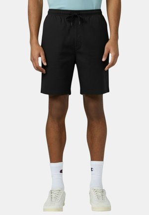 Uomo con pantaloncini neri con coulisse, calzini sportivi bianchi e scarpe da ginnastica bianche, in piedi con le braccia rilassate lungo i fianchi.