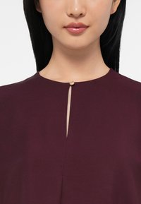 Woman with straight black hair wearing a plum-colored top featuring a small gold button and keyhole neckline detail.