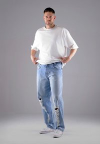 Weißes übergroßes T-Shirt, hellblaue distressed Jeans mit großen Rissen und weiße Sneakers mit roten Akzenten. Insgesamt ein schlichtes, lässiges Design.