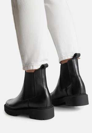 Czarne skórzane botki Chelsea z elastycznymi panelami po bokach, grubą czarną gumową podeszwą i teksturowanym wykończeniem. Wysokość do kostki, wyposażone w pętlę do zakładania.