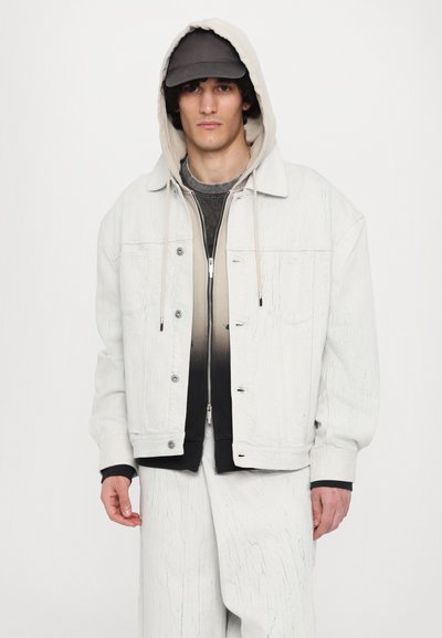 Chaqueta de mezclilla blanca con textura arrugada, botones plateados y capucha con cordón. Combinada con una camiseta en degradado de negro a gris por debajo.