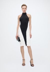 Γυναίκα με ασπρόμαυρο αμάνικο φόρεμα μήκους μέχρι το γόνατο, κρατώντας ένα μαύρο τσαντάκι χειρός, φορώντας μαύρα ψηλοτάκουνα σανδάλια, στέκεται μπροστά σε λευκό φόντο.