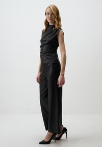 Zwart faux leren top met een hoge hals en kapmouwen, gecombineerd met zwarte high-waisted faux leren broek en puntige hoge hakken.