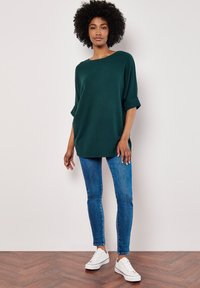 Dunkelgrünes, strukturiertes, übergroßes Oberteil mit aufgerollten Ärmeln, kombiniert mit blauen engen Jeans und weißen Sneakern auf einem braunen Fischgrätboden.