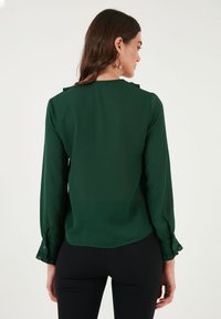 Dunkelgrüne Bluse mit langen Ärmeln, Rüschensäumen und glatter Textur. Der Rücken hat einen hohen Kragen und dezente Details an den Schulternähten.