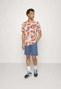 Κοντομάνικο πουκάμισο με φλοράλ μοτίβα σε κρεμ χρώμα με πορτοκαλί και μπλε σχέδια, συνδυασμένο με μπλε σορτς. Λευκές κάλτσες και σκούρα αθλητικά παπούτσια ολοκληρώνουν την εμφάνιση.