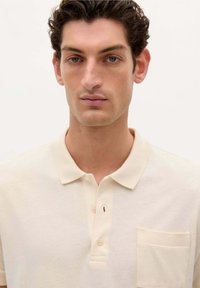 Camisa polo de color beige claro con cuello, tapeta de tres botones y un pequeño bolsillo en el pecho con un sutil detalle de costura. Textura de tela suave.