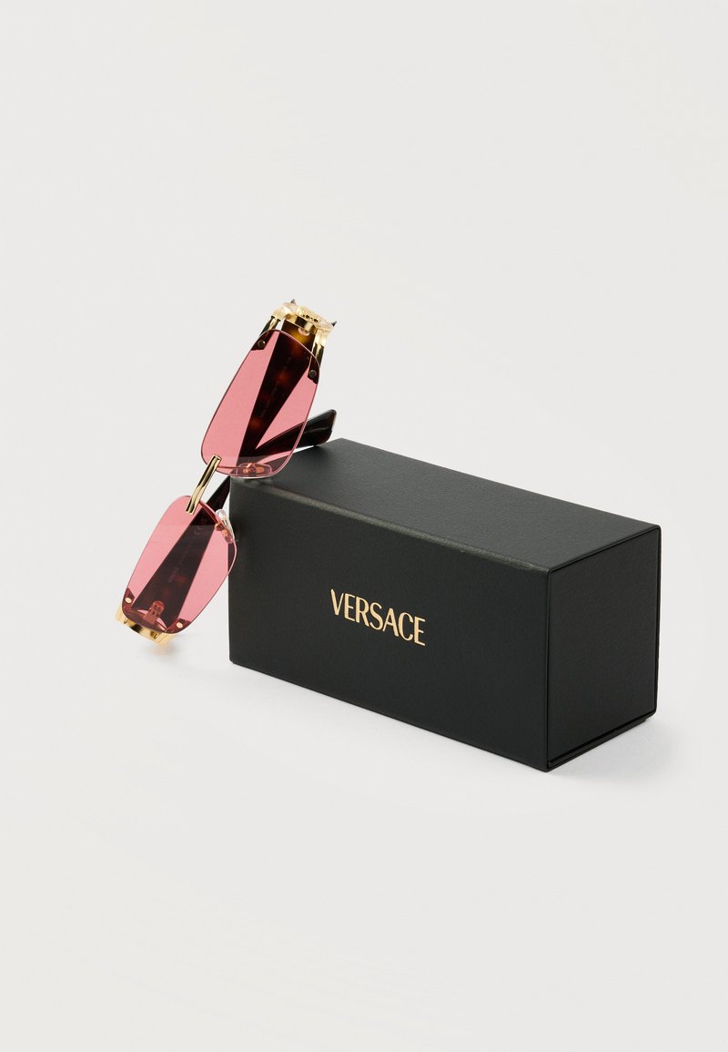Lunettes de soleil rectangulaires à verres roses avec des accents dorés posées sur une boîte noire Versace sur un fond blanc.