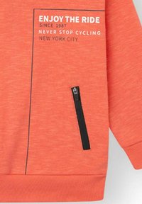 Sudadera naranja con un bolsillo negro con cremallera y texto blanco: "DISFRUTA DEL VIAJE", "DESDE 1987" y "NUNCA DEJES DE CICLAR, NUEVA YORK".