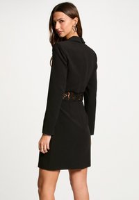 Donna con lunghi capelli castani che indossa un vestito nero a maniche lunghe con una cintura di pizzo, girata di spalle e guarda oltre la spalla.